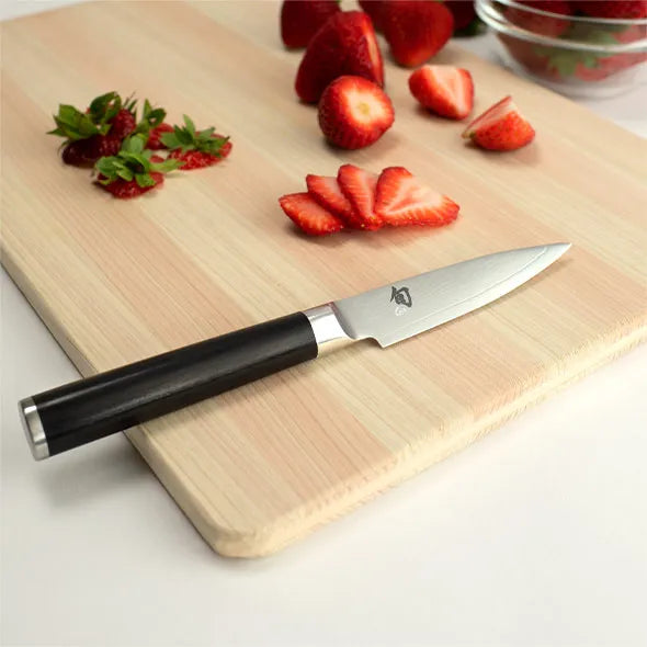 A Shun Classic paring knife with a black handle and silver blade resting on a light wood cutting board, with sliced strawberries and whole strawberries placed around it on a white surface.